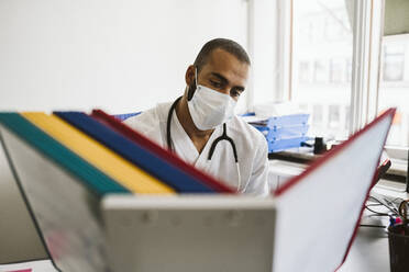 Männlicher Mitarbeiter des Gesundheitswesens beim Lesen von Akten in einer medizinischen Klinik während COVID-19 - MASF24041