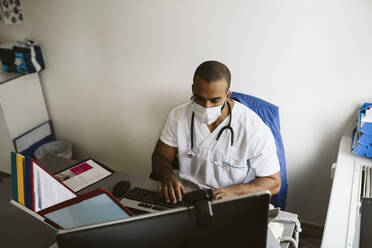 Male healthcare worker wearing face mask using computer in medical clinic - MASF24042