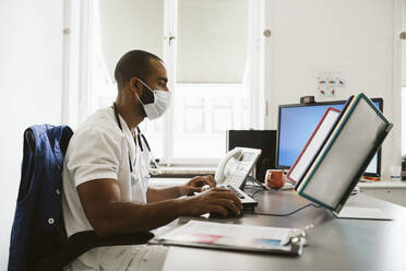 Männlicher Arzt, der am Schreibtisch sitzend einen Computer benutzt, während COVID-19 - MASF24043