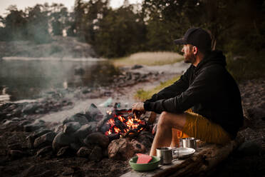 Side view of mature man sitting by campfire while looking away during sunset - MASF24045