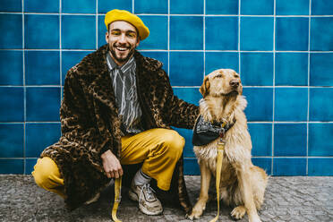 Lächelnder Mann in voller Länge, der neben einem Hund an einer blau gekachelten Wand kauert - MASF24053