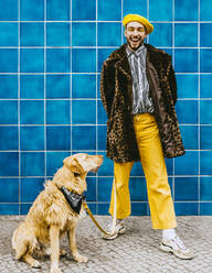 Full length of happy young man with dog standing against blue tiled wall - MASF24056