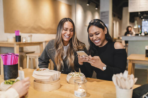 Lächelnde Freundinnen benutzen ihr Smartphone, während sie im Restaurant sitzen - MASF24106
