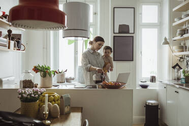 Weibliche Fachkraft gießt Kaffee ein, während sie ihren kleinen Sohn in der Küche trägt - MASF24114