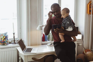 Male entrepreneur using smart phone while carrying baby boy at home office - MASF24118