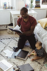 Male entrepreneur kneeling while reading diary by son at home - MASF24147
