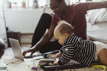 Männliches Kleinkind spielt mit Spielzeug, während der Vater im Schlafzimmer einen Laptop benutzt - MASF24148