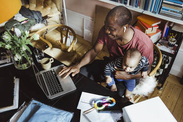 Smiling businessman using laptop while sitting with male toddler at home office - MASF24171