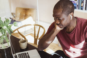 Besorgter männlicher Unternehmer mit Hand am Kinn, der auf seinen Laptop schaut, während er von seinem Heimbüro aus arbeitet - MASF24175