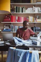 Male entrepreneur writing while examining shirt at home office - MASF24177