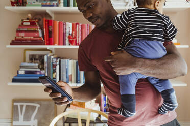 Father using smart phone while carrying baby boy at home - MASF24179