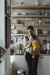 Mutter schreibt eine SMS auf ihrem Smartphone, während sie ihren kleinen Sohn in der Küche trägt - MASF24197