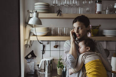 Mutter telefoniert mit Smartphone, während sie ihr Kleinkind in der Küche trägt - MASF24198