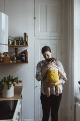 Mother carrying baby boy while standing in kitchen - MASF24199