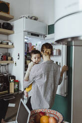 Rückansicht einer Mutter, die einen kleinen Jungen trägt, während sie den Kühlschrank in der Küche öffnet - MASF24206