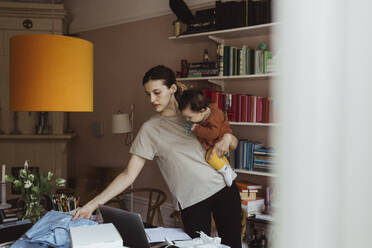 Geschäftsfrau schaut auf ihr Hemd, während sie ein männliches Kleinkind im Wohnzimmer trägt - MASF24232