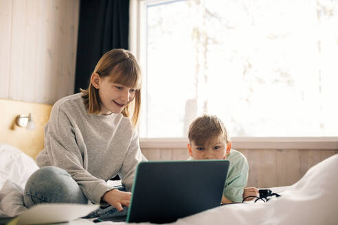 Lächelndes Mädchen mit Bruder, der einen Laptop im Schlafzimmer zu Hause benutzt - MASF24247