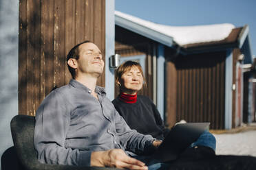 Männliche und weibliche Berufstätige sitzen mit geschlossenen Augen vor einem Ferienhaus an einem sonnigen Tag - MASF24249