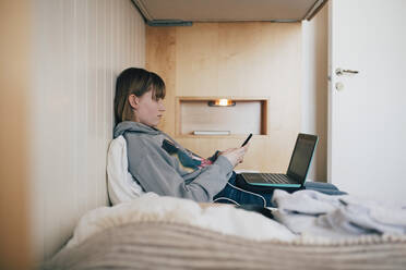 Teenager-Mädchen sitzt mit Laptop und benutzt ein Smartphone im Ferienhaus - MASF24256