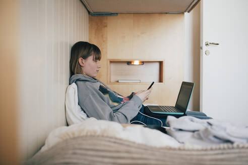 Teenage girl sitting with laptop using smart phone in vacation home - MASF24256