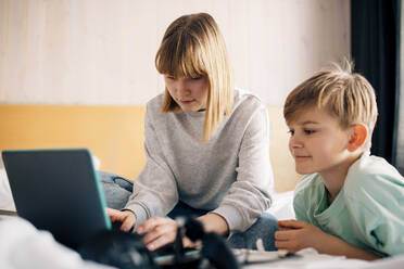 Teenager-Mädchen, das einen Laptop benutzt, während es neben seinem Bruder auf dem Bett zu Hause sitzt - MASF24279