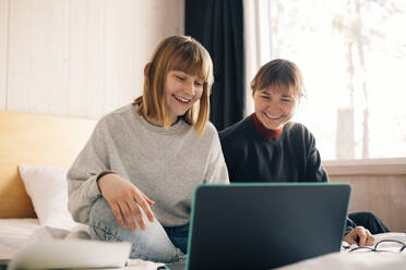 Lächelnde Mutter und Tochter sitzen mit Laptop beim Hausunterricht im Schlafzimmer - MASF24281