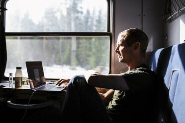 Geschäftsmann, der einen Laptop benutzt, während er im Zug am Fenster sitzt - MASF24292