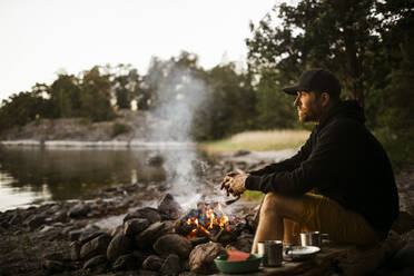 Seitenansicht eines nachdenklichen Mannes, der wegschaut, während er am Lagerfeuer am Seeufer sitzt - MASF24303