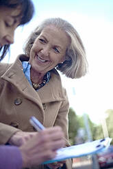 Lächelnde ältere Frau schaut auf eine Frau, die einen Vertrag auf einem Klemmbrett unterzeichnet - AJOF01443