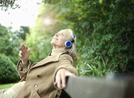 Lächelnde ältere Frau schaut auf, während sie auf einer Bank im öffentlichen Park Musik hört - AJOF01449