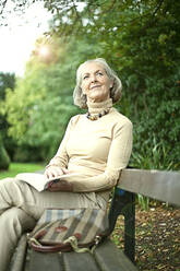 Ältere Frau mit Buch schaut weg, während sie auf einer Bank im Park sitzt - AJOF01450