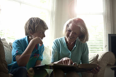 Lächelnde Frau zeigt ihrem Enkel ein Fotoalbum, während sie zu Hause auf dem Sofa sitzt - AJOF01451