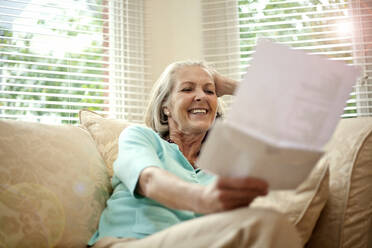 Glückliche ältere Frau liest einen Brief, während sie auf dem Sofa im Wohnzimmer sitzt - AJOF01452