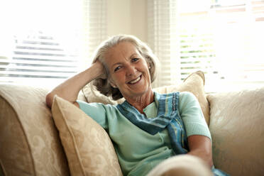 Lächelnde ältere Frau mit dem Kopf in der Hand auf dem Sofa zu Hause sitzend - AJOF01453