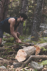 Young man cutting fallen tree trunk with axe in forest - JAQF00673