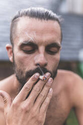 Shirtless young man smoking marijuana joint - JAQF00676