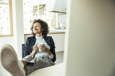 Female entrepreneur holding coffee cup while relaxing at office - UUF23439