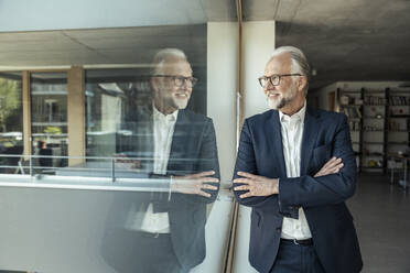 Smiling businessman with arms crossed leaning on glass window at office - UUF23447