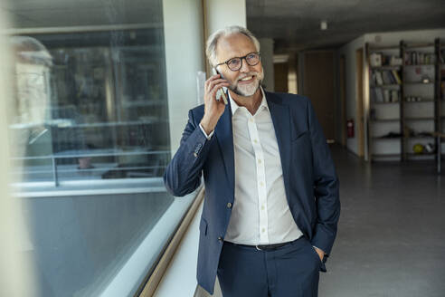 Geschäftsmann, der in der Nähe eines Glasfensters im Büro mit seinem Smartphone telefoniert - UUF23452