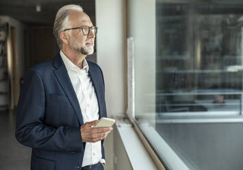 Geschäftsmann schaut durch ein Fenster und hält ein Smartphone im Büro - UUF23455