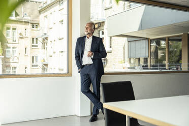 Männlicher Berufstätiger, der eine Kaffeetasse hält und sich an ein Glasfenster im Büro lehnt - UUF23460