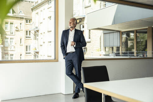 Männlicher Berufstätiger, der eine Kaffeetasse hält und sich an ein Glasfenster im Büro lehnt - UUF23460