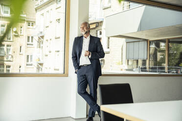 Nachdenklicher männlicher Berufstätiger, der sich an ein Glasfenster im Büro lehnt - UUF23461