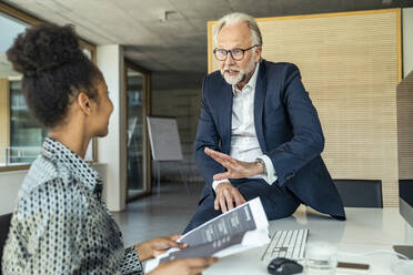 Businessman discussing with female colleague while sitting on desk at office - UUF23469