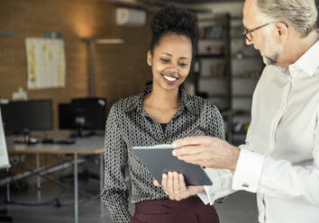 Männlicher Unternehmer zeigt einer lächelnden Kollegin im Büro ein digitales Tablet - UUF23477