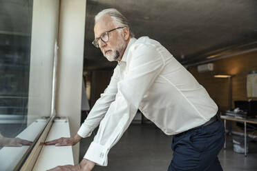 Ernster Geschäftsmann, der durch das Fenster schaut, während er sich an sein Büro lehnt - UUF23484