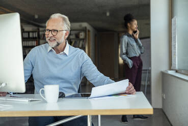 Male professional sitting at desk while businesswoman talking in background - UUF23487