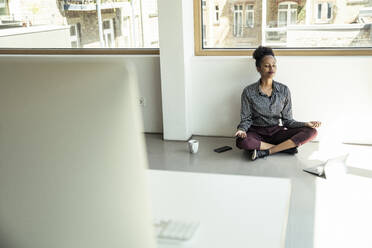 Junge Geschäftsfrau meditiert, während sie im Büro auf dem Boden sitzt - UUF23495
