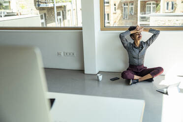 Lächelnde weibliche Fachkraft mit Kopf in den Händen sitzt auf dem Boden im Büro - UUF23496