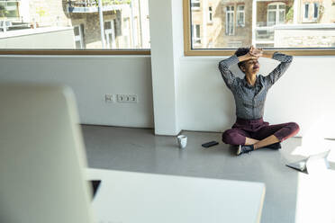 Smiling female professional with head in hands sitting on floor at office - UUF23496